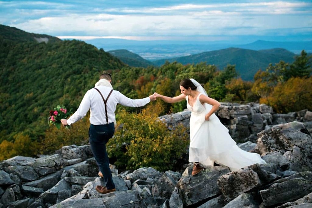 A couple holds hands and navigates rocks in wedding attire during an adventure elopement in Virginia