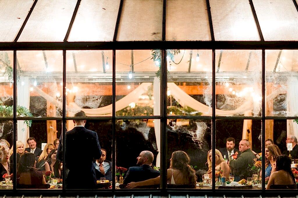 A couple listens to a toast in the greenhouse at Avonlea Farms. The photographer is outside the greenhouse looking in