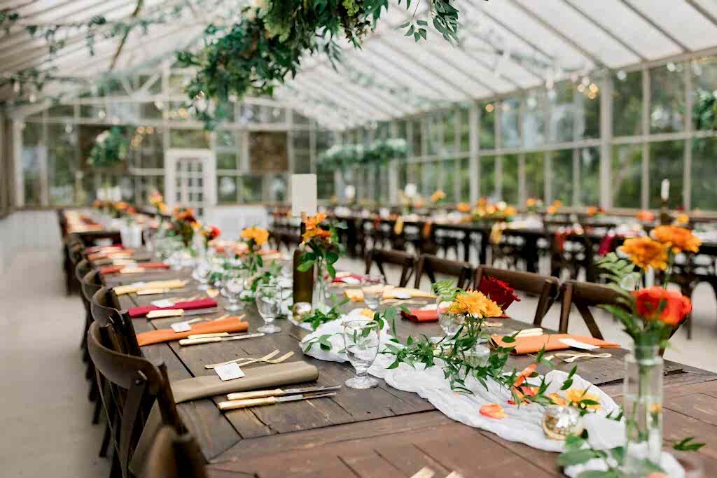 A tablescape in a greenhouse wedding venue shows lots of colorful flowers and greenery